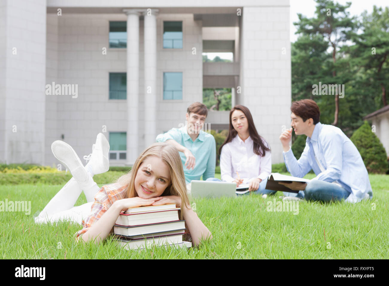 Internationale Schüler in der Schule mit dem Kopf auf den Bauch auf einer Wiese liegend ruhte auf einem Stapel Bücher mit anderen Studenten zusammen hinter ihr Stockfoto