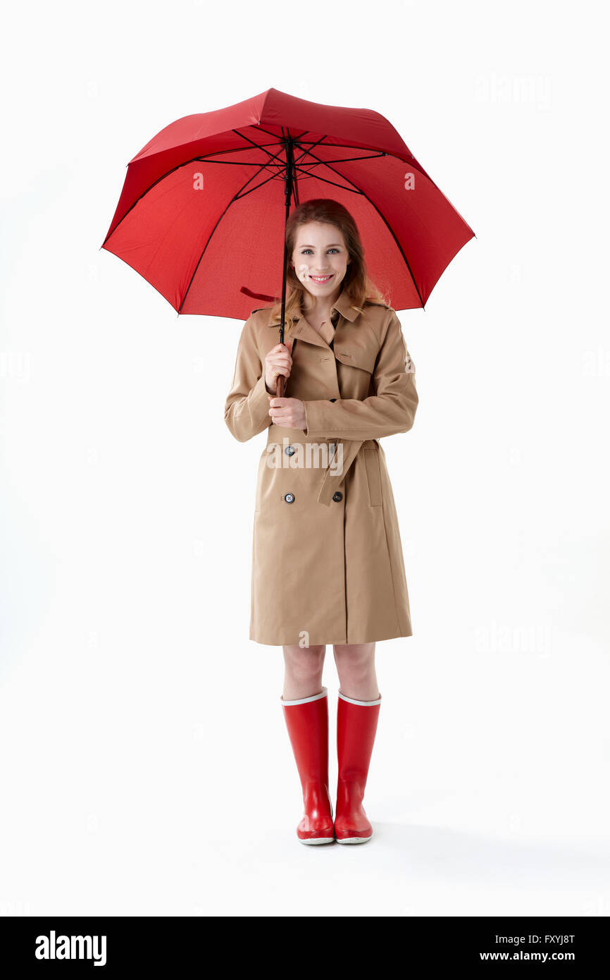 Frau im Trenchcoat und roten Regen Stiefel halten einen roten Regenschirm und Stand mit einem Lächeln Stockfoto