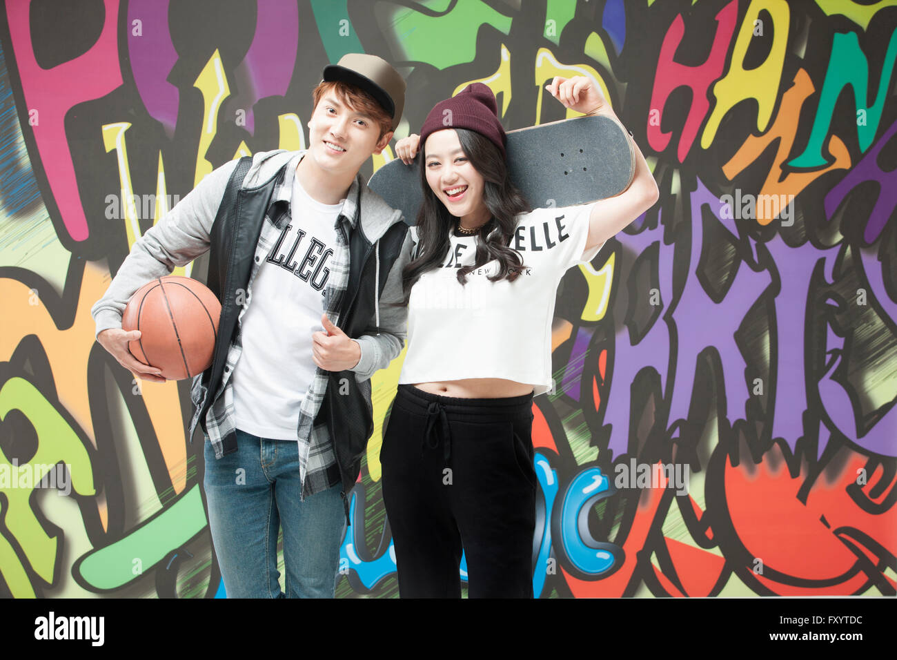 Junge, lächelnde paar im Hip-Hop-Stil hält ein Basketball- und ein Skateboard gegen Graffiti-Kunst Stockfoto