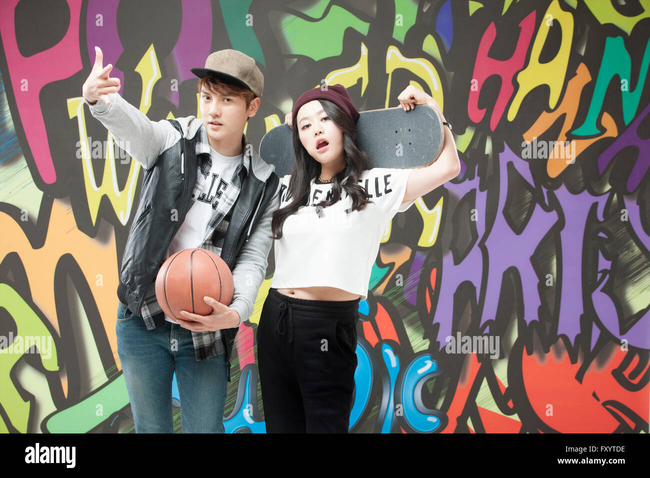 Junges Paar im Hip-Hop-Stil hält ein Basketball- und ein Skateboard gegen Graffiti-Kunst Stockfoto