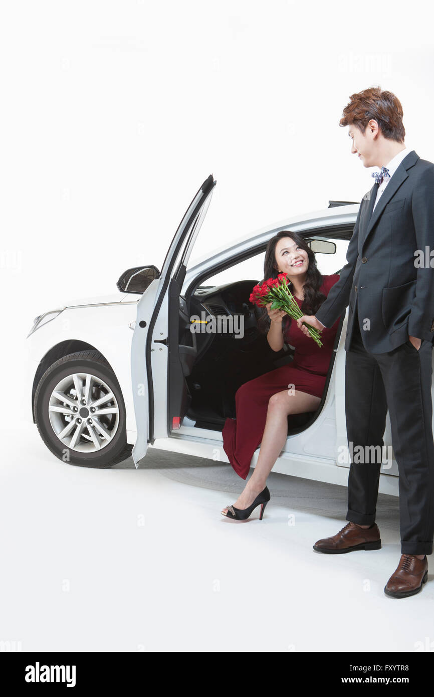 Junge lächelnde Frau sitzt in Oepn Auto ein Strauß roter Rosen von einem Mann in Anzug vor dem Fahrzeug steht unter Stockfoto