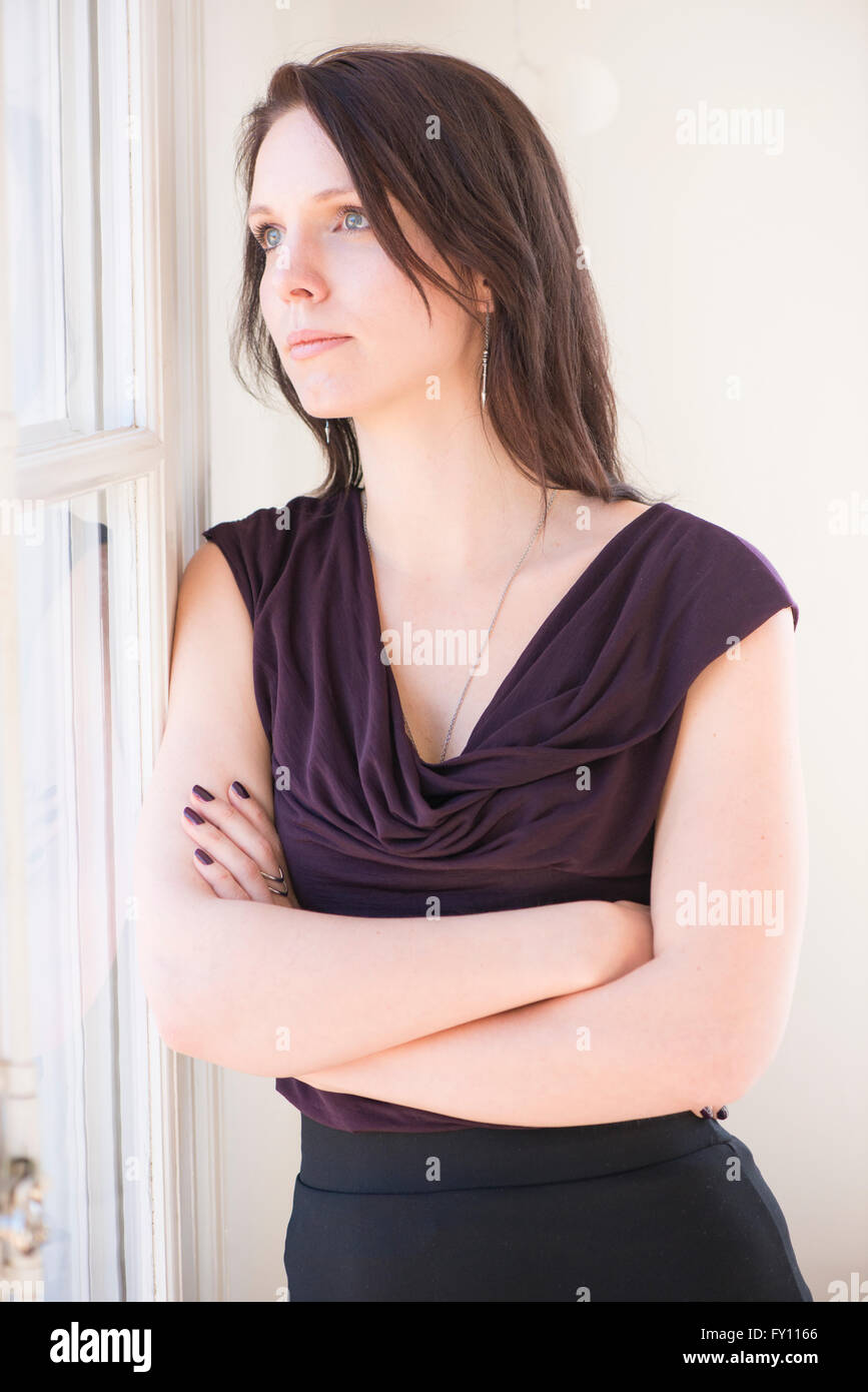 Frau im schwarzen Kleid ein Fenster stand, wegschauen. Sie hat verschränkten Armen und einem nachdenklichen Ausdruck. Stockfoto