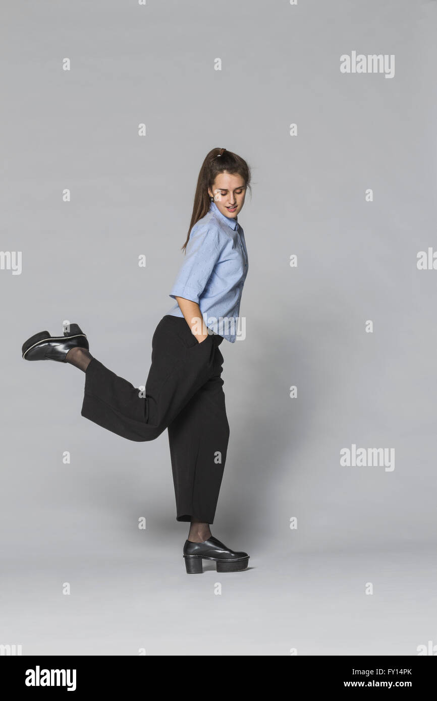 Voller Länge Frau stehen auf einem Bein mit Händen in den Taschen vor grauem Hintergrund Stockfoto