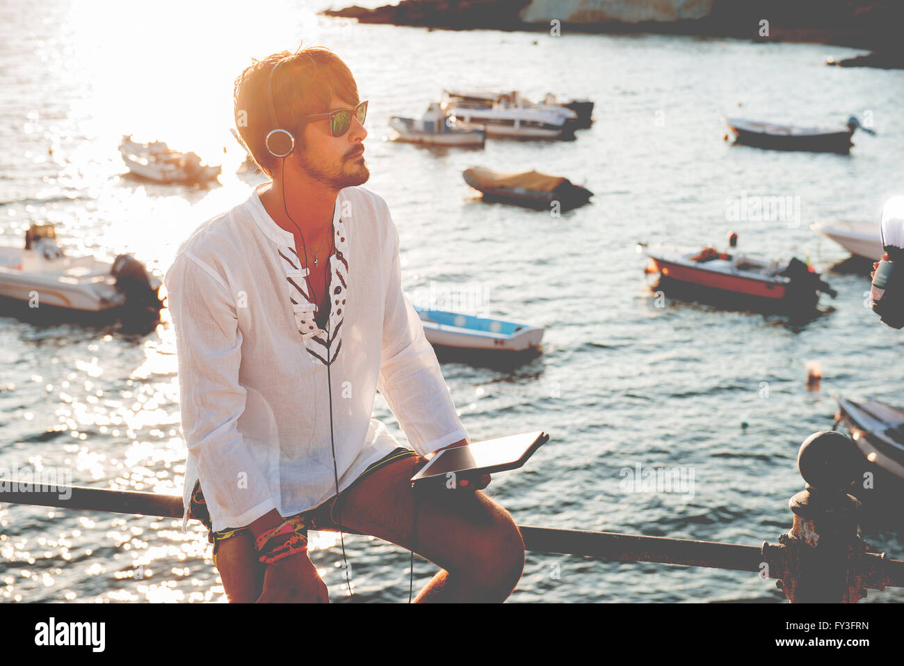 junger Mann Musik hören mit Kopfhörern aus Tablet in einer Ferien-Destination, sitzt ein kleiner Hafen Stockfoto