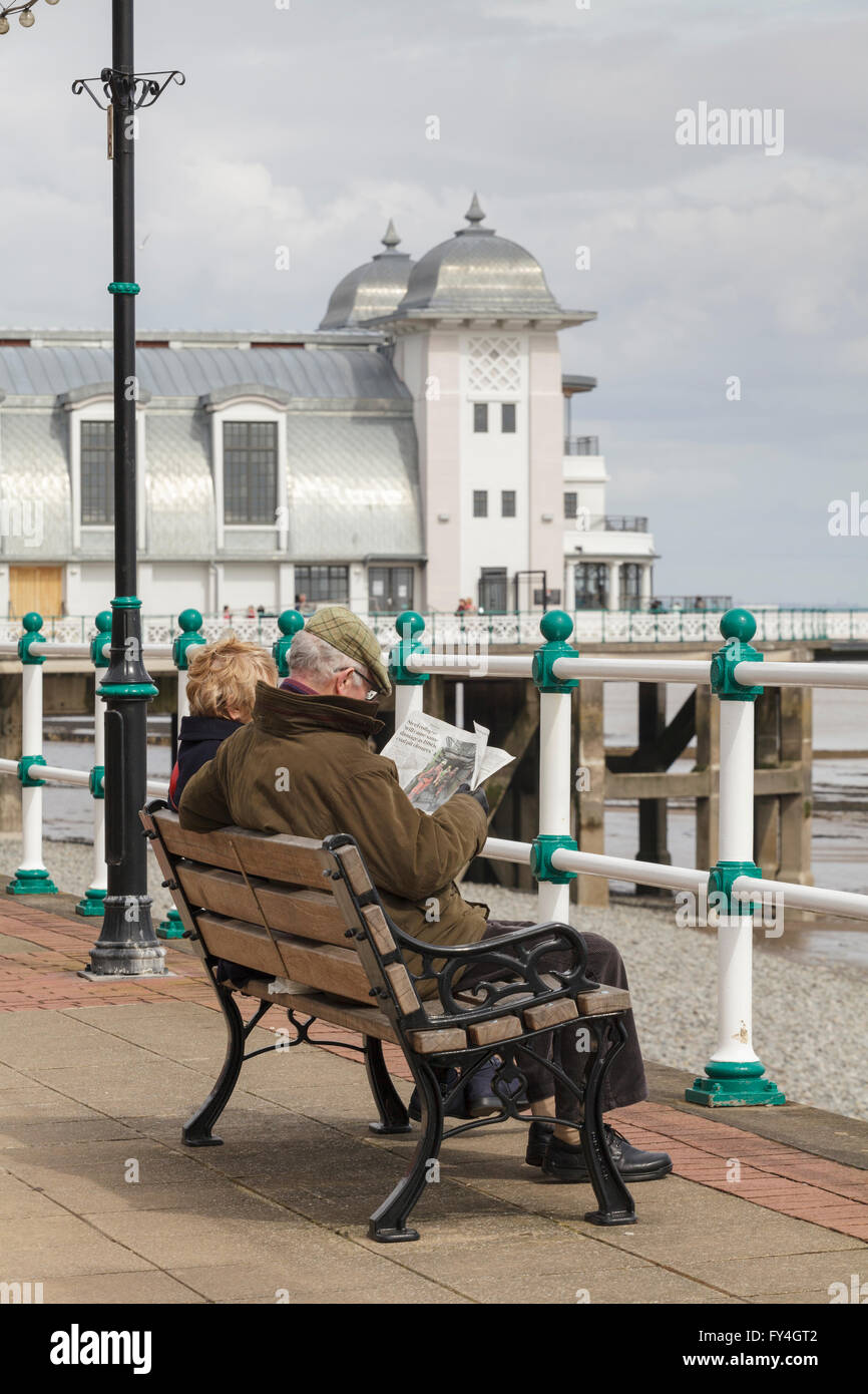 Ein älteres Ehepaar sitzt auf einer Bank und liest eine Zeitung in Penarth Pier. Stockfoto