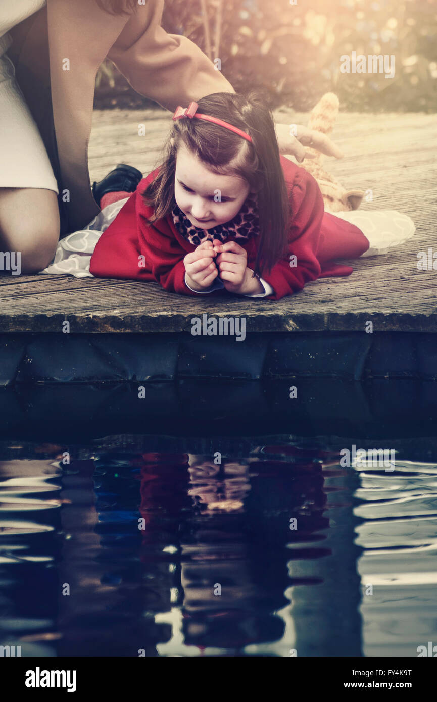 kleines Mädchen mit ihrer Mutter sitzt neben dem Teich blickte auf Wasserreflexion Stockfoto