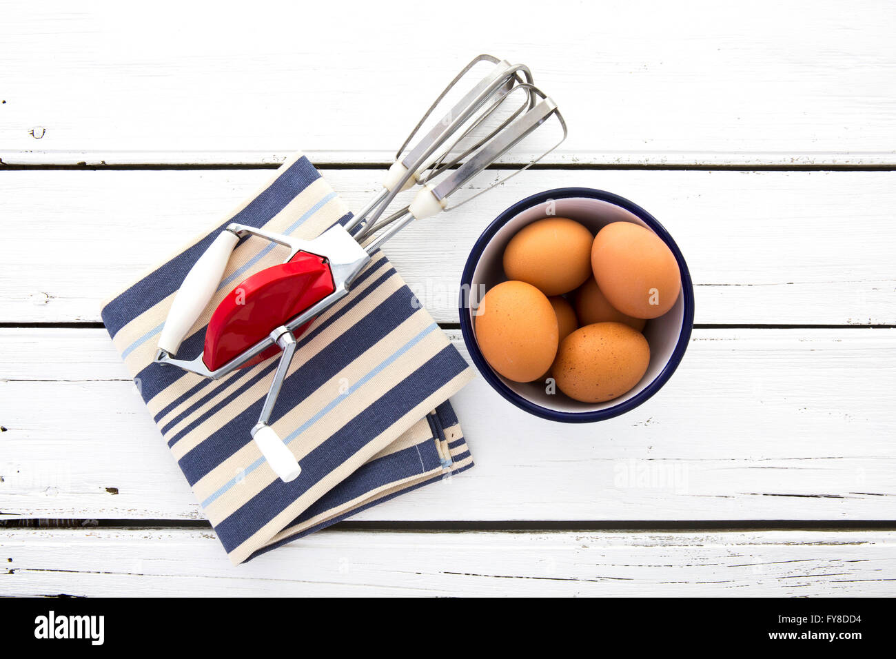 Vintage Metall Hand Schneebesen mit roten Körper Schuss auf Retro-weißen hölzernen Hintergrund mit blau-weiß gestreiften Serviette und Schüssel Eier Stockfoto