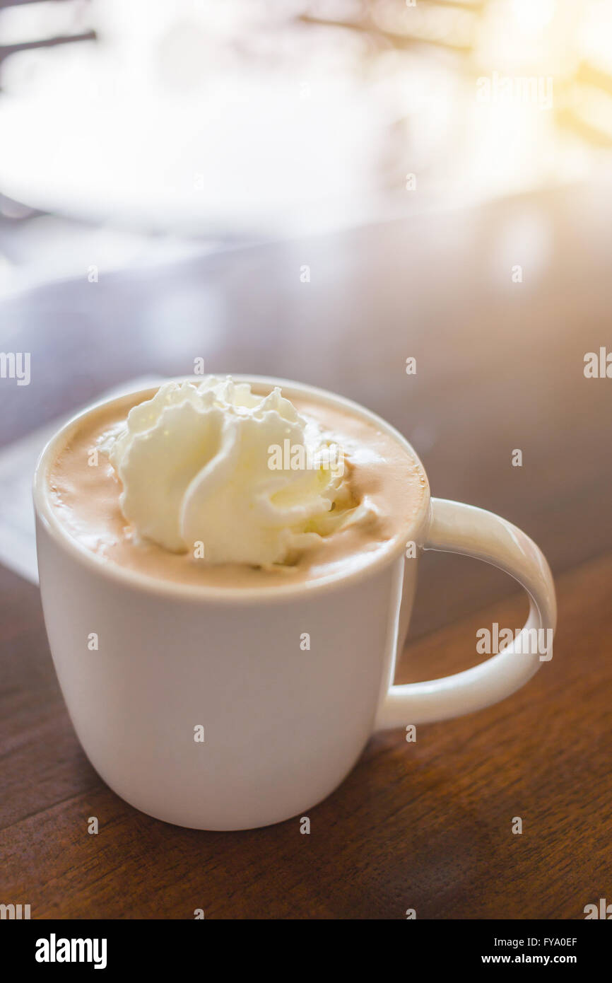 Fokus auf heißen Mokkakaffee mit Milch-Sahne in weißen Tasse auf Holztisch und helles Licht in der Nähe Stockfoto