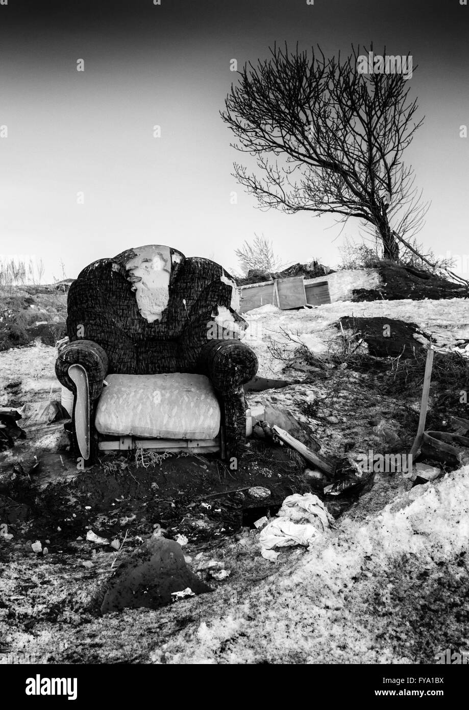 Verlassene Einzelsessel in einem sich verschlechternden Zustand umgeben von Detritus als Baum beugt sich hinüber in den Hintergrund. Monochrome bla Stockfoto