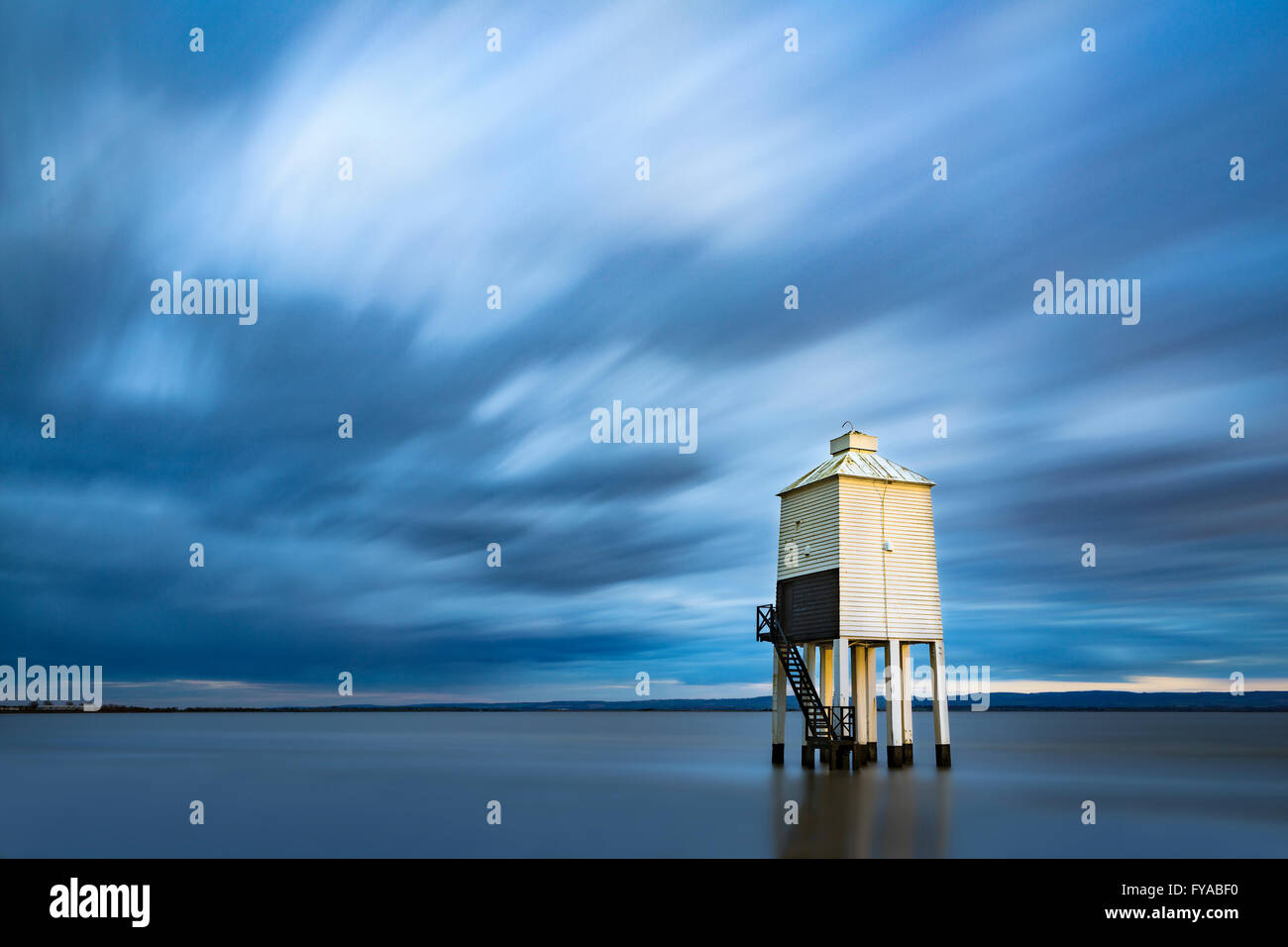 Die niedrige Leuchtturm am Burnham-on-Sea wird durch eine Welle von Sonnenlicht bei Sonnenuntergang beleuchtet. Eine Langzeitbelichtung verwischt die Wolken und die steigende Flut. Stockfoto