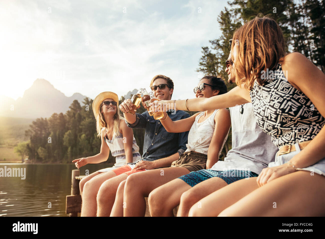 Porträt der jungen Menschen sitzen auf einem Steg und Toasten Bier an einem sonnigen Tag. Glückliche junge Männer und Frauen hängen am See Stockfoto