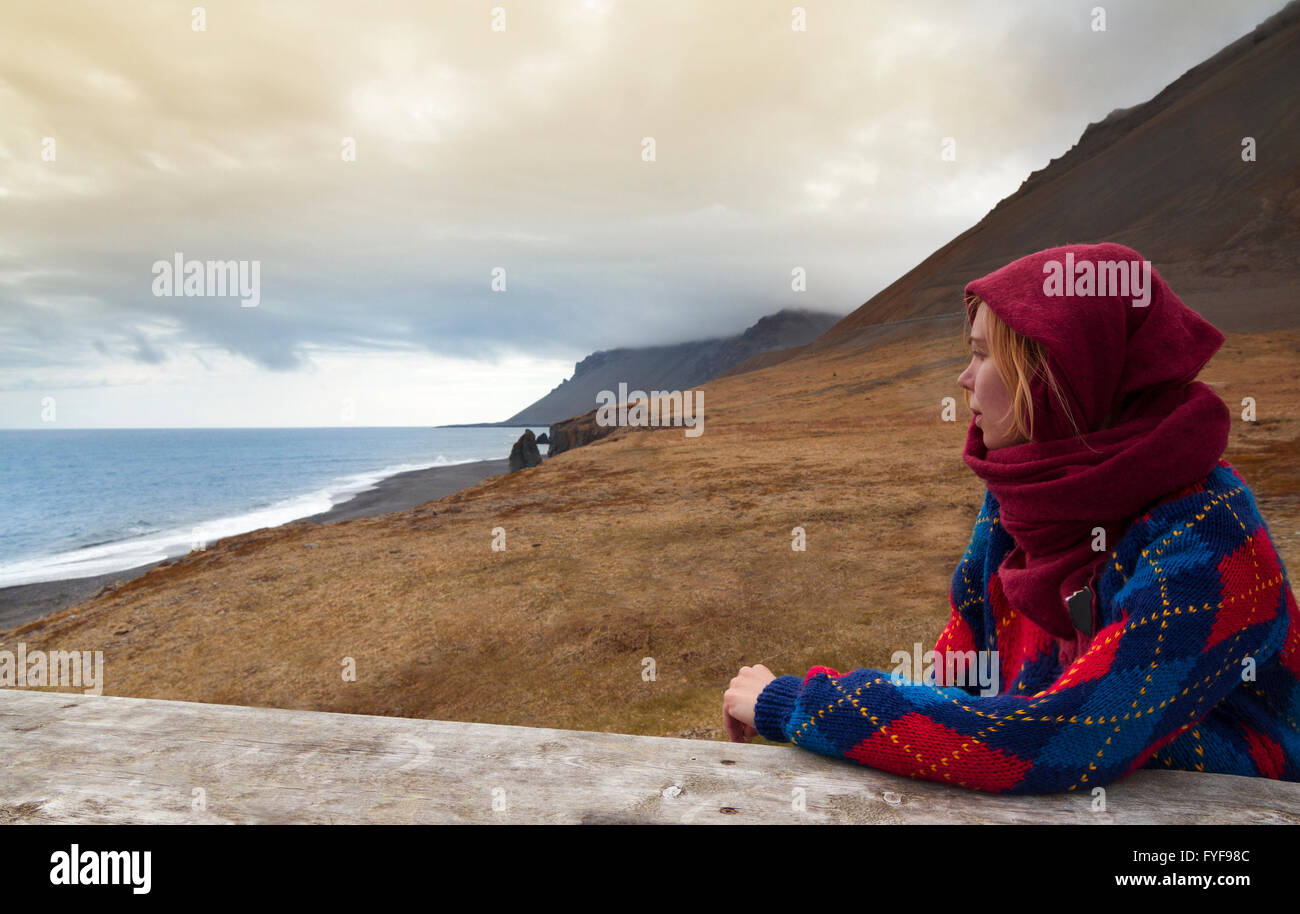 Horizontale Foto einer jungen blonden Frau trägt einen Schal und einen Pullover und mit Blick auf eine wunderschöne Küstenlinie in Island Stockfoto
