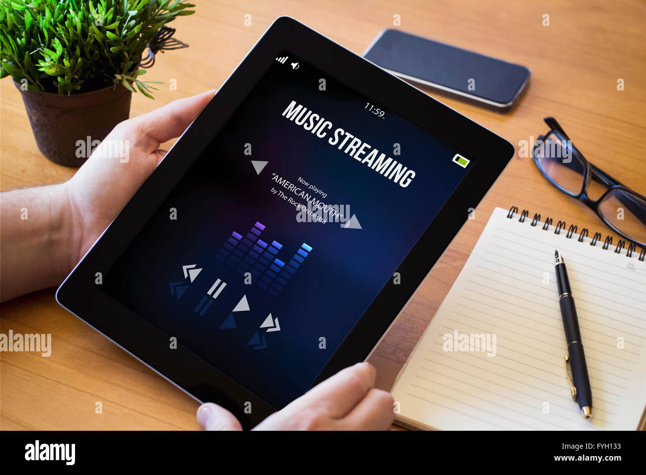 Hände eines Mannes holding Tablet mit Musik-streaming über eine hölzerne Workspace-Tabelle. Alle Bildschirm-Grafiken bestehen. Stockfoto