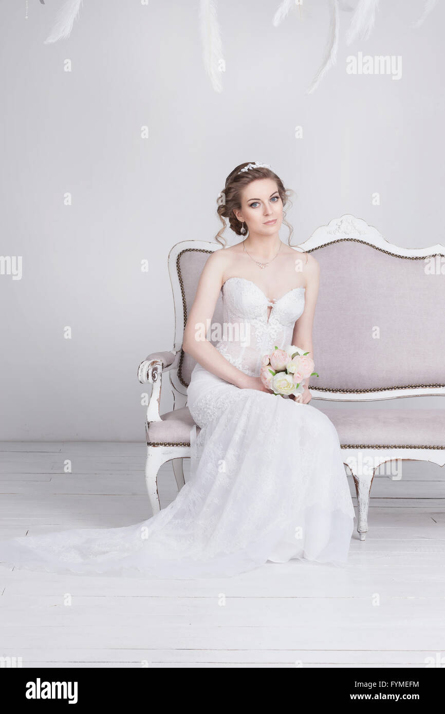 Schöne junge Braut im Hochzeitskleid edler Spitze. Sie sitzt auf einem weißen Sofa Vintage. Sie ist eine elegante und schlanke, sie hat p Stockfoto