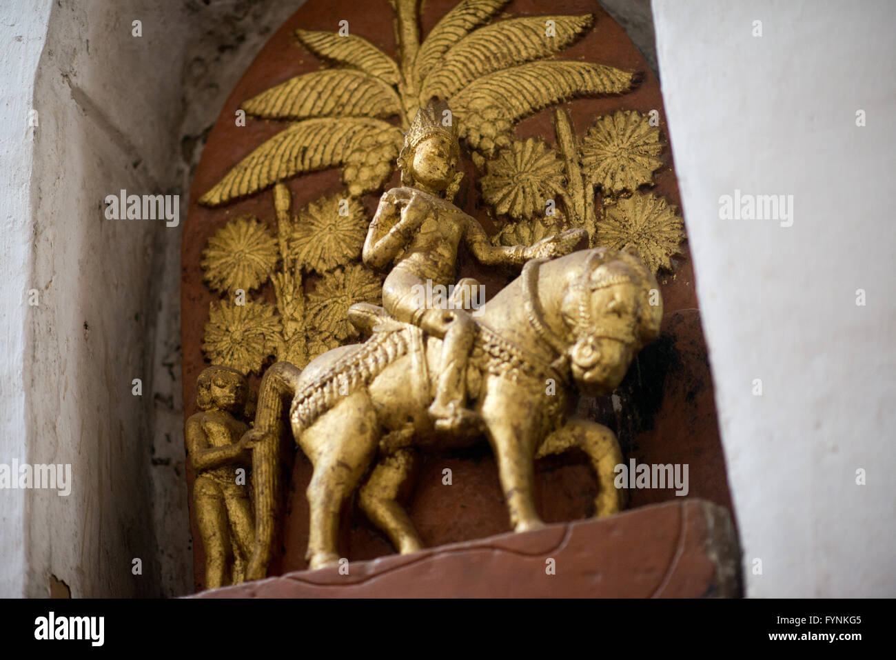 Ananda Temple Buddha Reitpferd Bagan Myanmar // BAGAN, Myanmar — Eine kleine goldene Buddha-Figur, die auf einem Pferd mit Palmen im Hintergrund reitet, befindet sich in einer Nische hoch auf einer Innenwand des Ananda Tempels und stellt einen Teil der umfangreichen narrativen Kunstwerke dar, die das komplexe Korridorsystem des Tempels schmücken. Diese kleineren skulpturalen Szenen stellen wahrscheinlich Episoden aus den Jataka-Geschichten oder anderen buddhistischen Schriften dar, was zur Rolle des Tempels als Zentrum für visuelle buddhistische Erziehung und Erzählkunst beiträgt. Der Tempel beherbergt über 1.500 Steinskulpturen und glasierte Tafeln mit Abbildungen Stockfoto