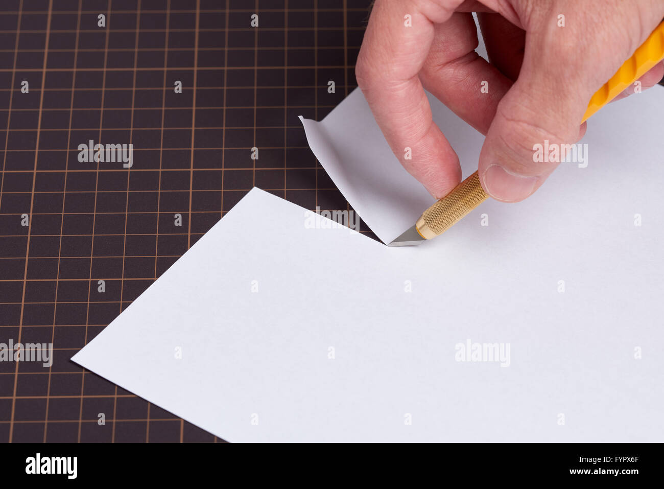 Eine Hand mit einem Messer mit einem gelben Griff schneiden ein Stück weißes Papier auf eine Schneidematte. Stockfoto