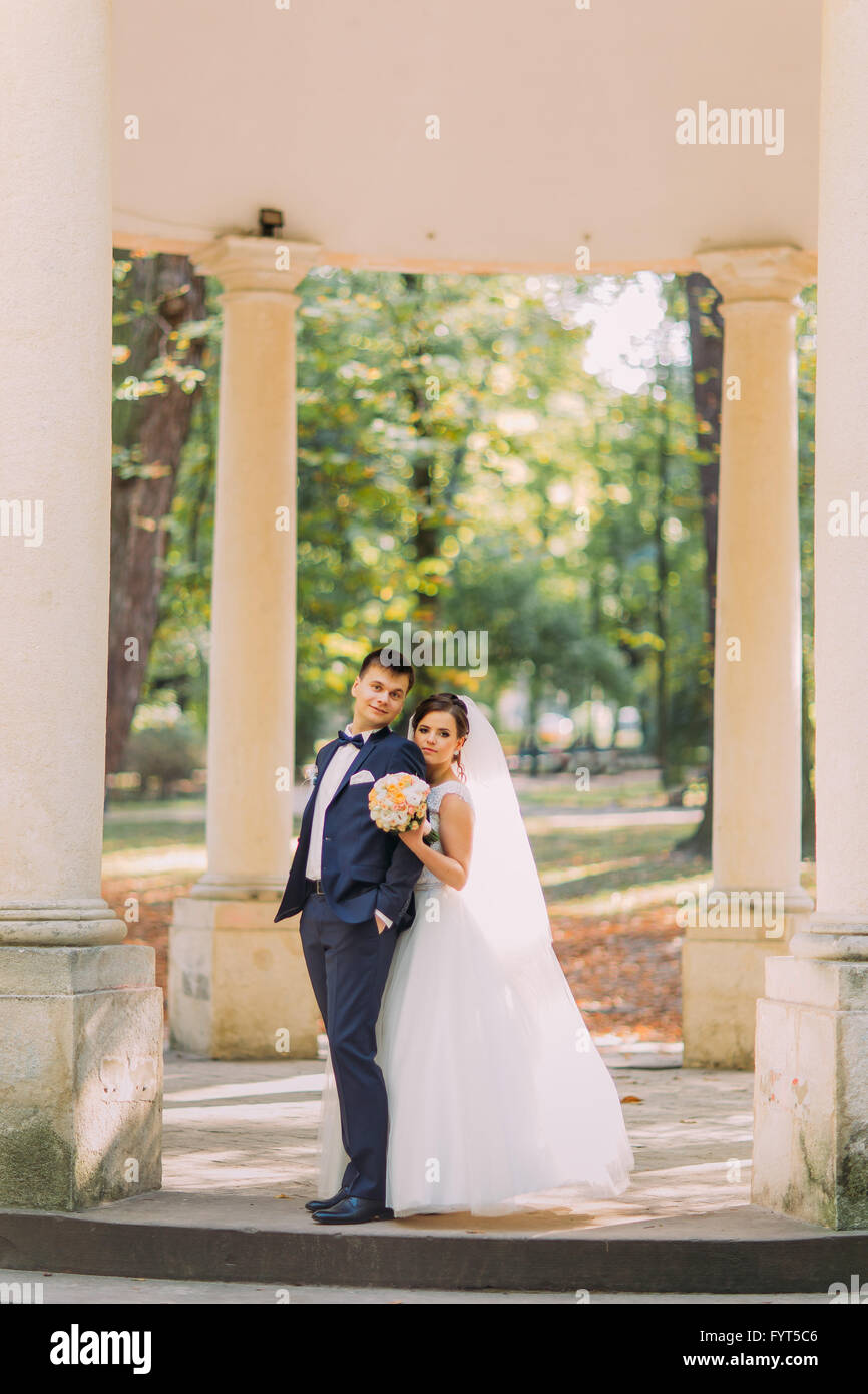 Schöne Braut umarmt Bräutigam von hinten vor dem Hintergrund der Gebäude mit Säulen Stockfoto