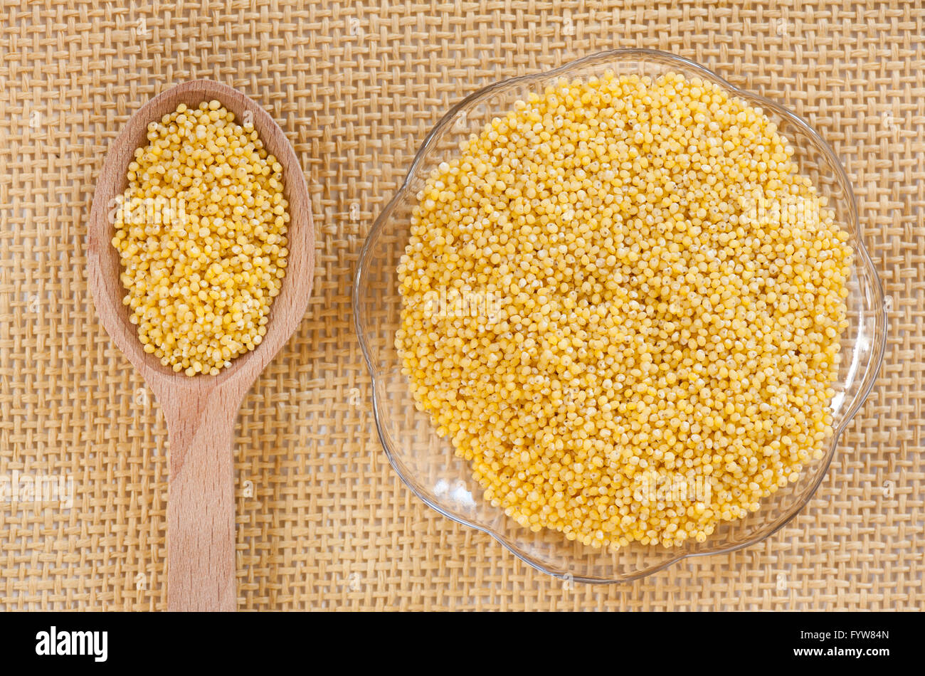 Gelbe Hirse geschältes Getreide auf Glas Untertasse und Holzlöffel auf Matte, gesunde Grundnahrungsmittel Rohprodukt, Teil der Samen liegen. Stockfoto