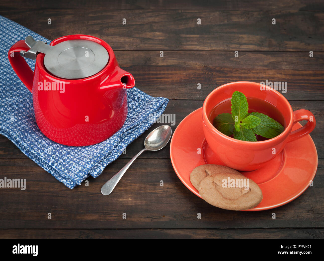 Rote Teekanne auf Teatowel, orange Teetasse mit Untertasse, Cookies, Minze und Löffel auf Holztisch mit textfreiraum gelegt. Stockfoto