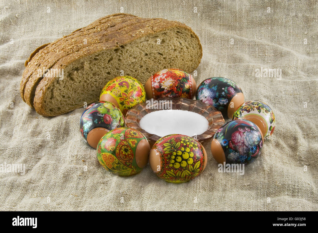 Viele frische ländliche dekorierte Ostereier liegend in einem wattled Körbchen in der Nähe von frischem Brot und Salzstreuer auf ein Tuch aus einem ge Stockfoto