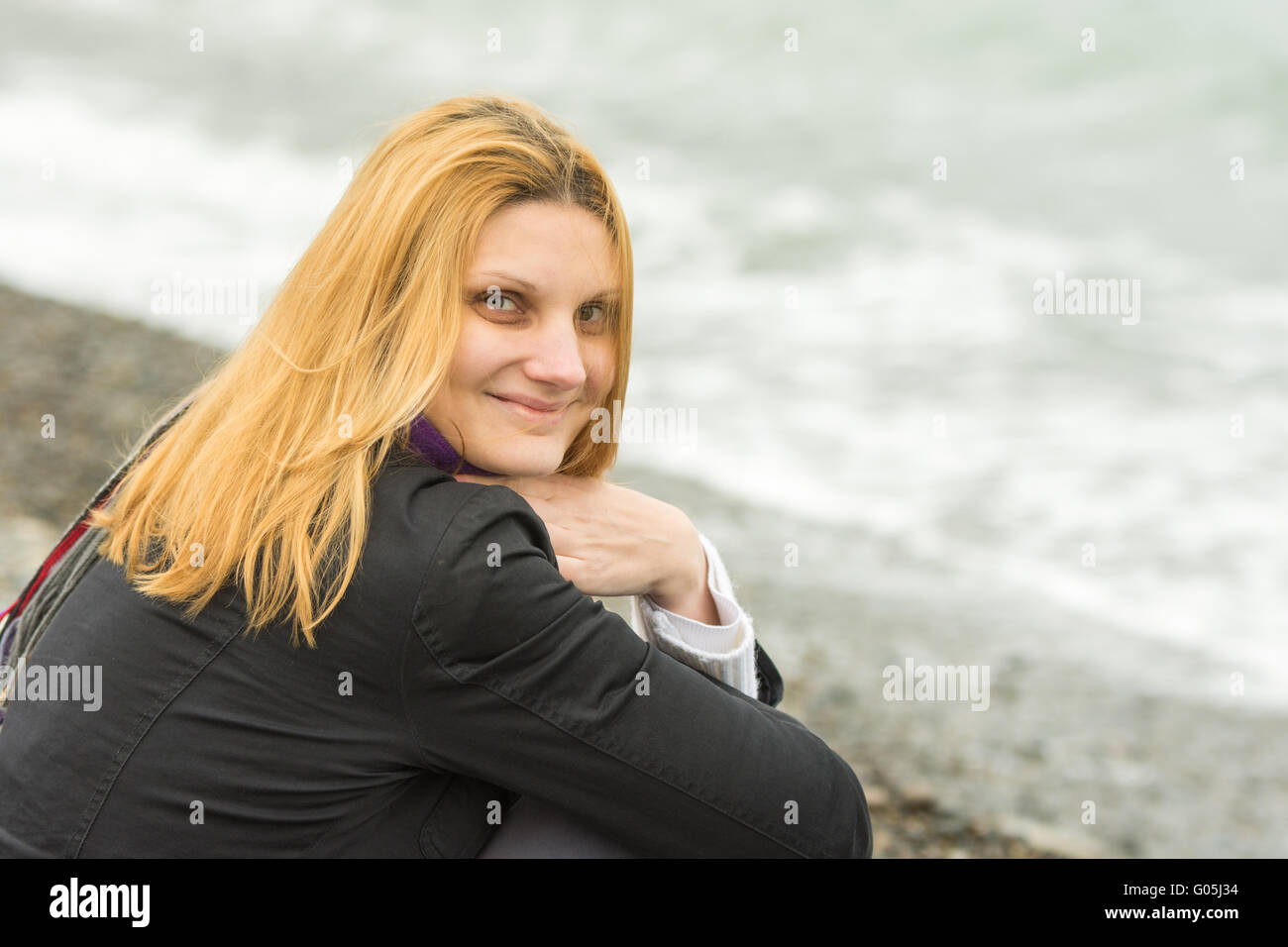 Porträt des Sitzens auf dem Strand lächelnde junge Frau in der kalten Jahreszeit Stockfoto
