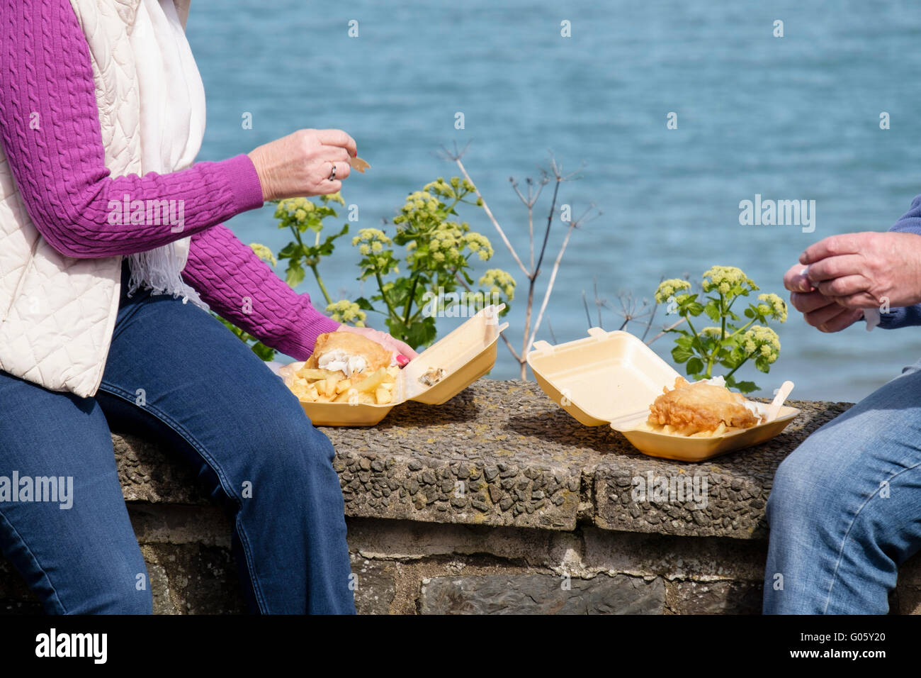 Zwei Personen Urlauber sitzen auf einem Meer wand Essen takeaway Fish und Chips aus Polystyrol Behälter in der Küstenstadt New Quay Wales UK Stockfoto