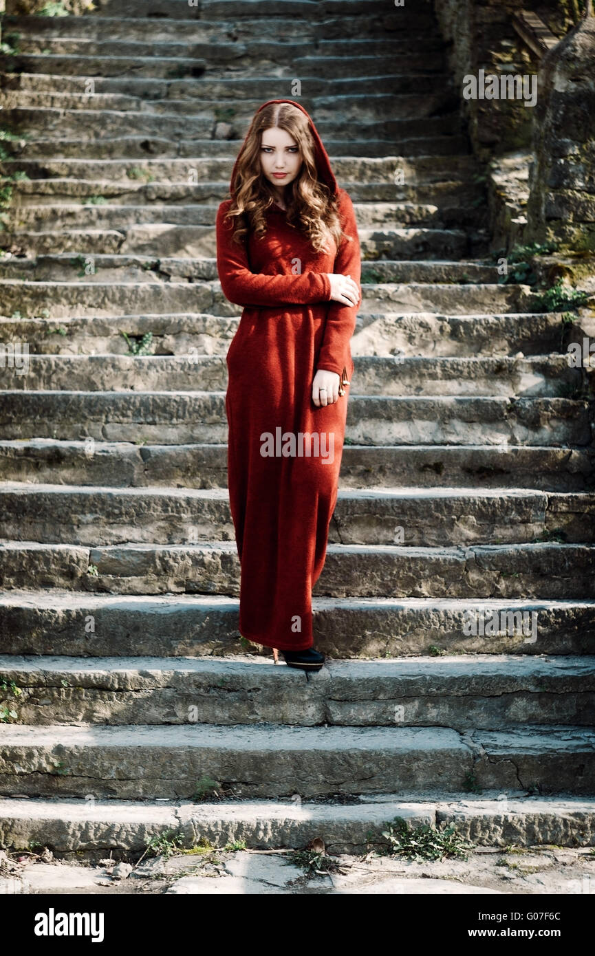 Porträt der schönen jungen Frau mit lockigem Haar im roten Kleid mit Kapuze sitzt auf alten Steintreppe Stockfoto
