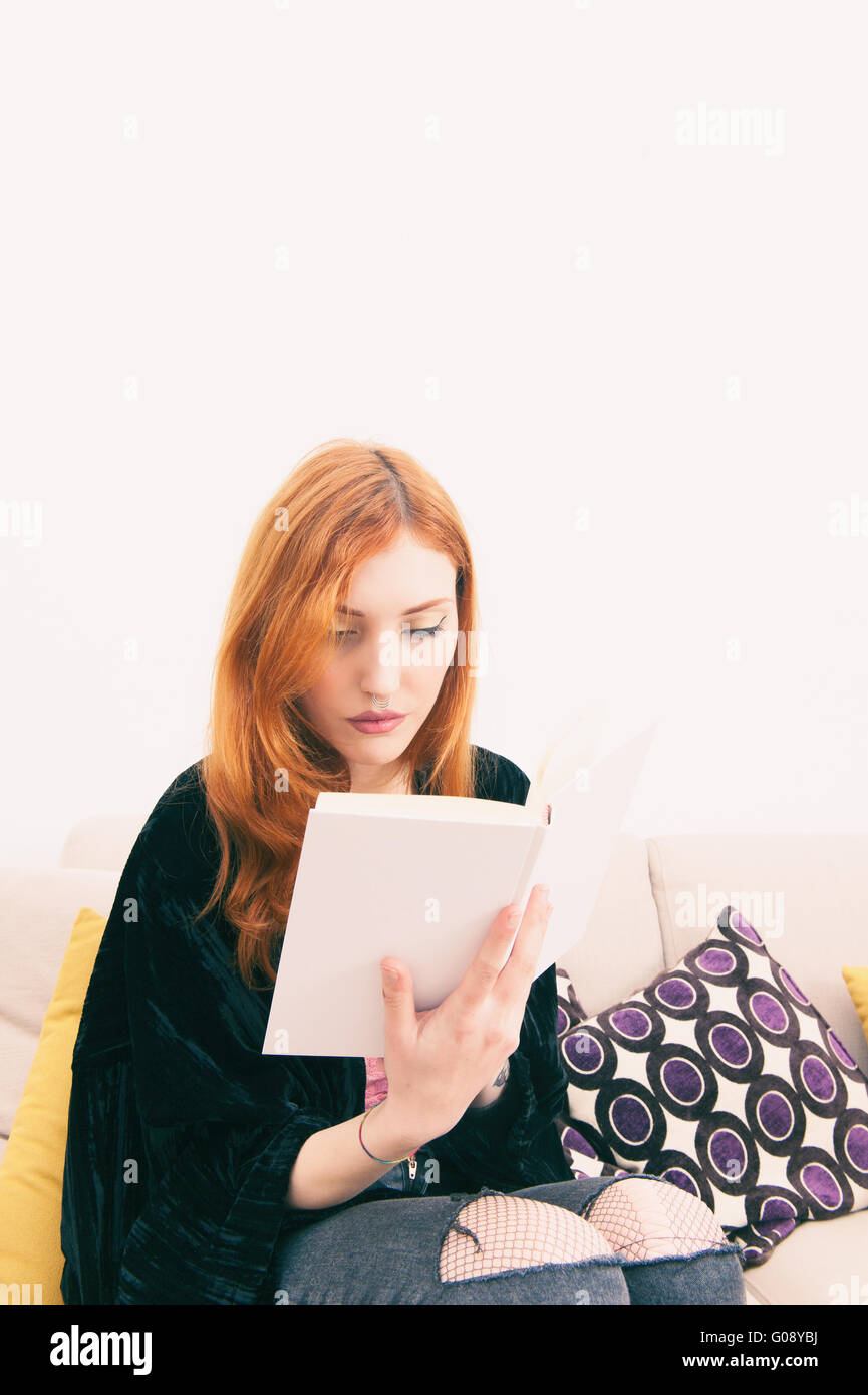 Junge rothaarige Frau liest ein Buch auf dem Sofa im Wohnzimmer sitzen Stockfoto