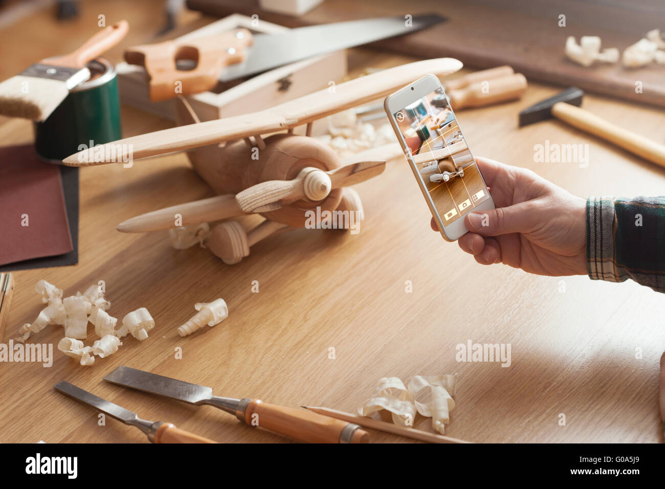 Mann fotografiert seine handgefertigten hölzernes Spielzeugflugzeug mit einem Smartphone auf einem Arbeitstisch Stockfoto