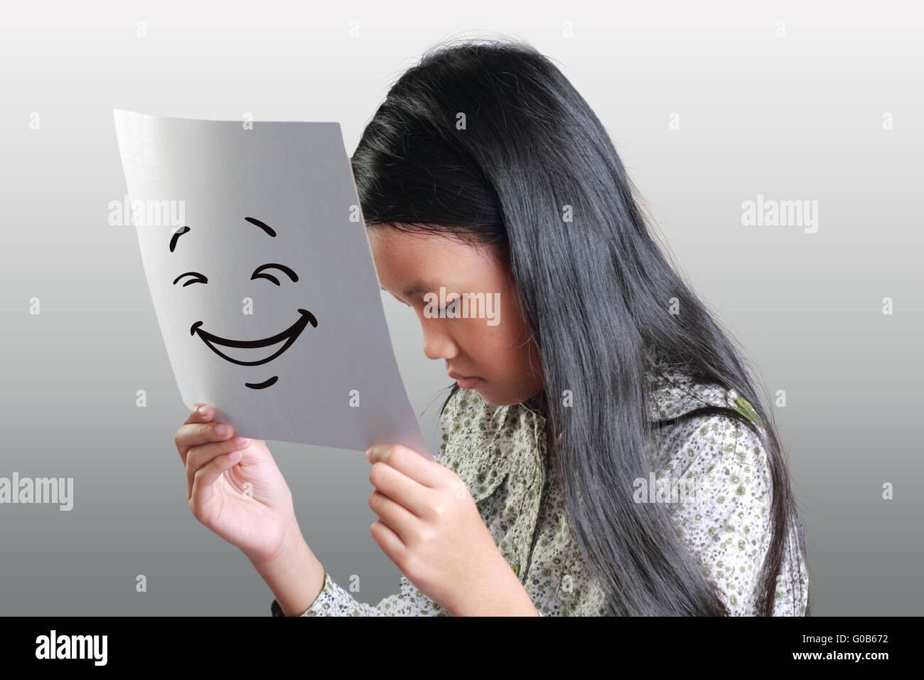 Traurige kleine Asiatin hält Weißbuch vor ihr glückliches Gesicht drauf gezogen Stockfoto