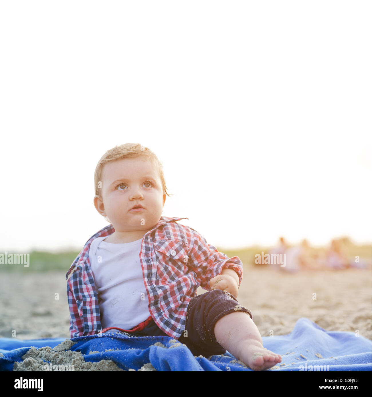 Gut aussehend neugieriges Kind sitzt auf Sand am Strand schaut zum Himmel empor am sonnigen Tag. Stockfoto