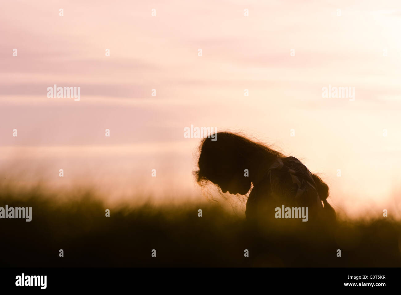 Mädchen im Profil vor lila Sonnenuntergang Silhouette. Kind mit langen Haaren sitzen lange Gras in der Abenddämmerung mit warmen Abendhimmel Stockfoto