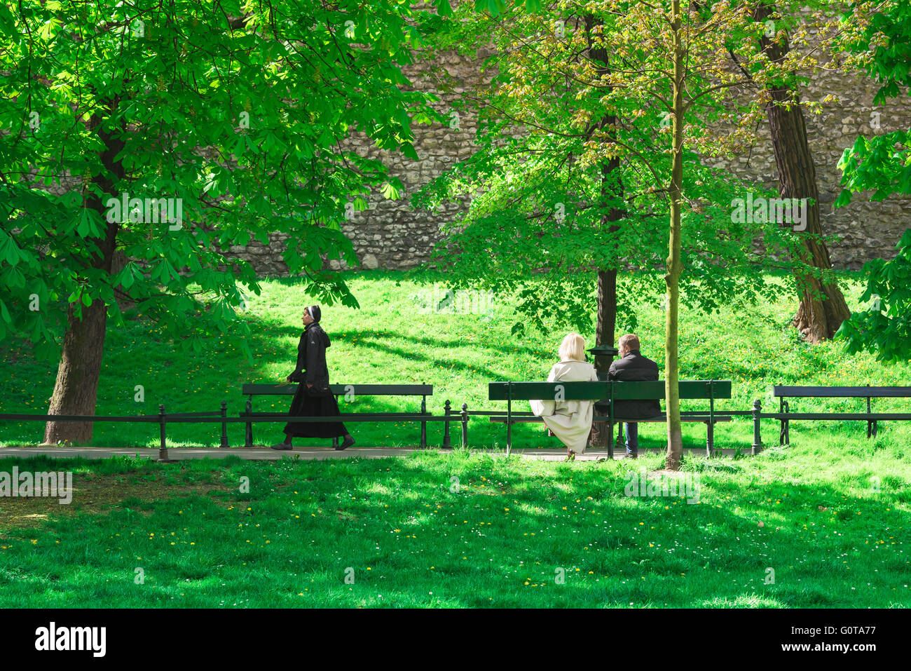 Dagegen, Blick auf eine Nonne, die an einem Paar vorbeirollt, das auf einer Bank im Planty Park, Krakow, Polen sitzt. Stockfoto