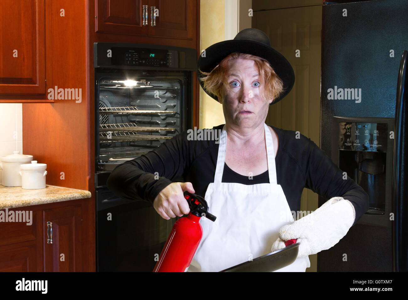 Frau mit Schürze und Pilger Hut Holding schwenken und Feuer Extenguisher mit verkohlten Gesicht und schockiert Ausdruck in Küche. Stockfoto