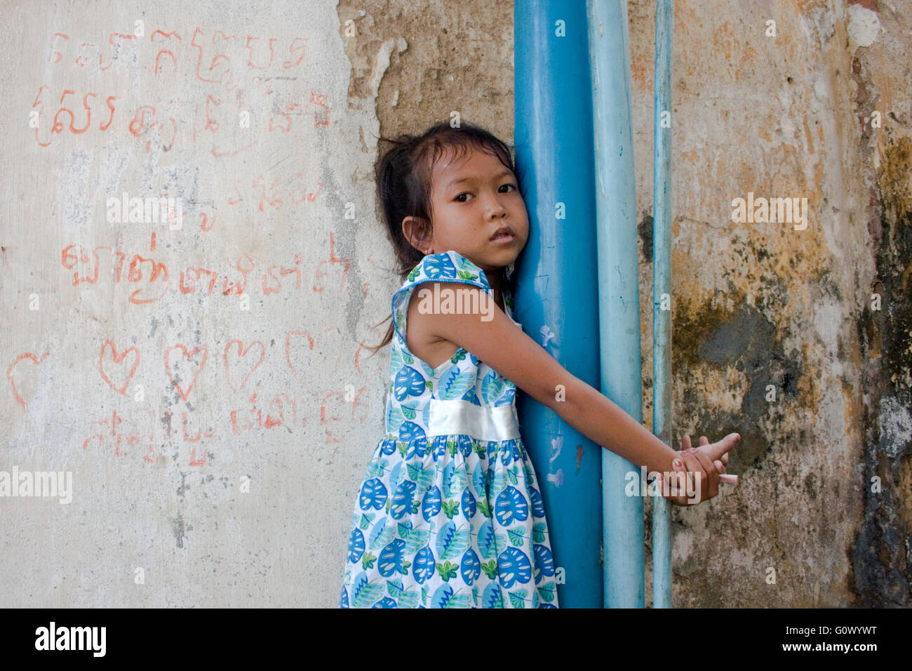 Ein junges Kleinkind Mädchen in Armut lebenden wickelt ihre Arme um Kunststoff Wasserleitungen in einem Slum in Kampong Cham, Kambodscha. Stockfoto