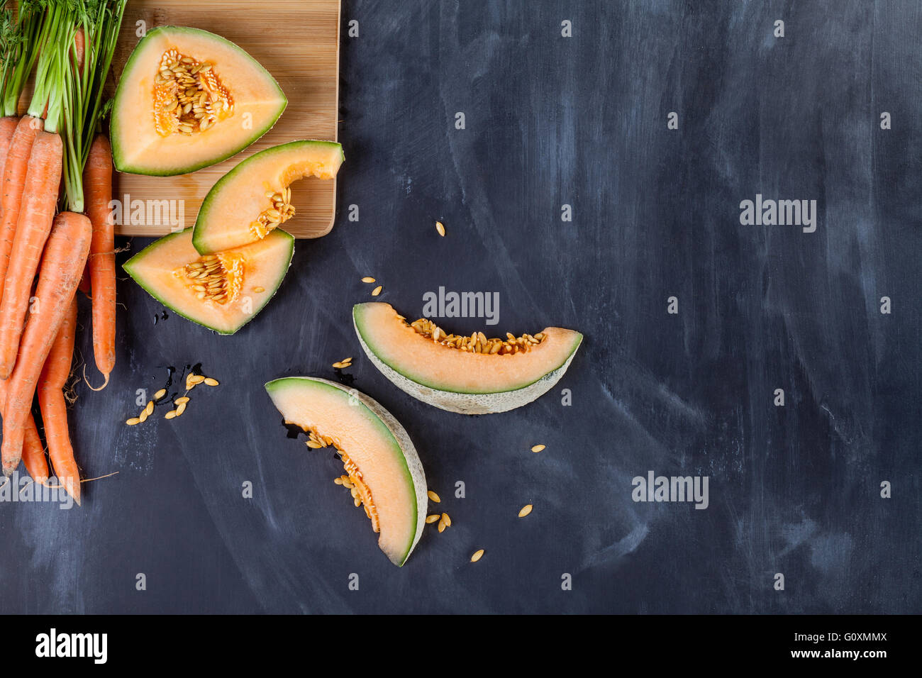 Scheiben von Melone und Haufen von Karotten auf Schneidebrett am schwarzen Brett Stockfoto