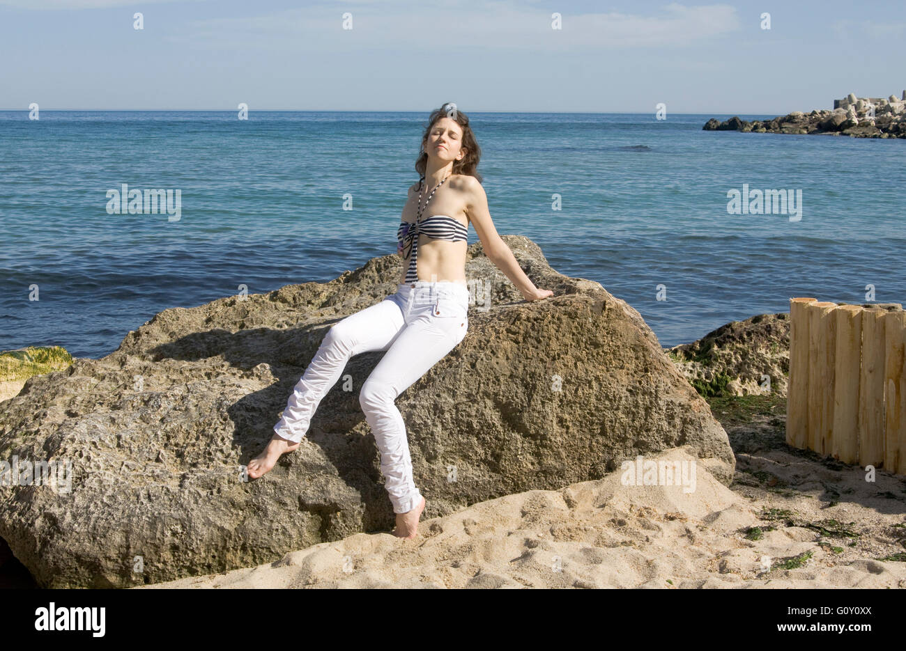 Junge europäische Frau in weißer Hose und Oberteil des Bikinis liegt am großen Stein am Strand. Stockfoto