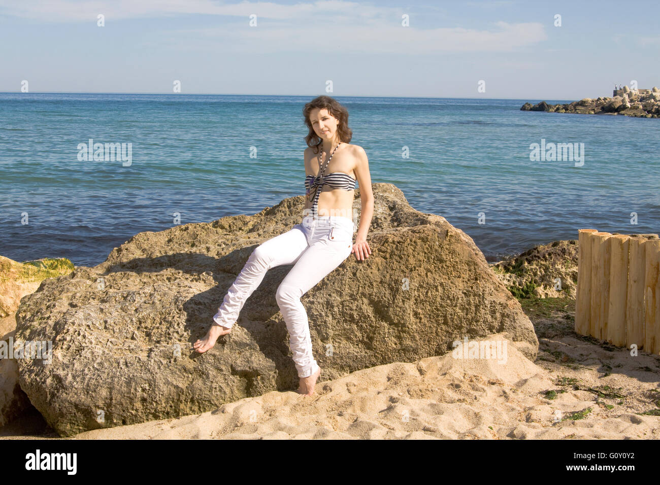 Europäische Frau mit braunen Haaren in weißer Hose und Oberteil des Bikinis liegt am großen Stein am Strand. Stockfoto