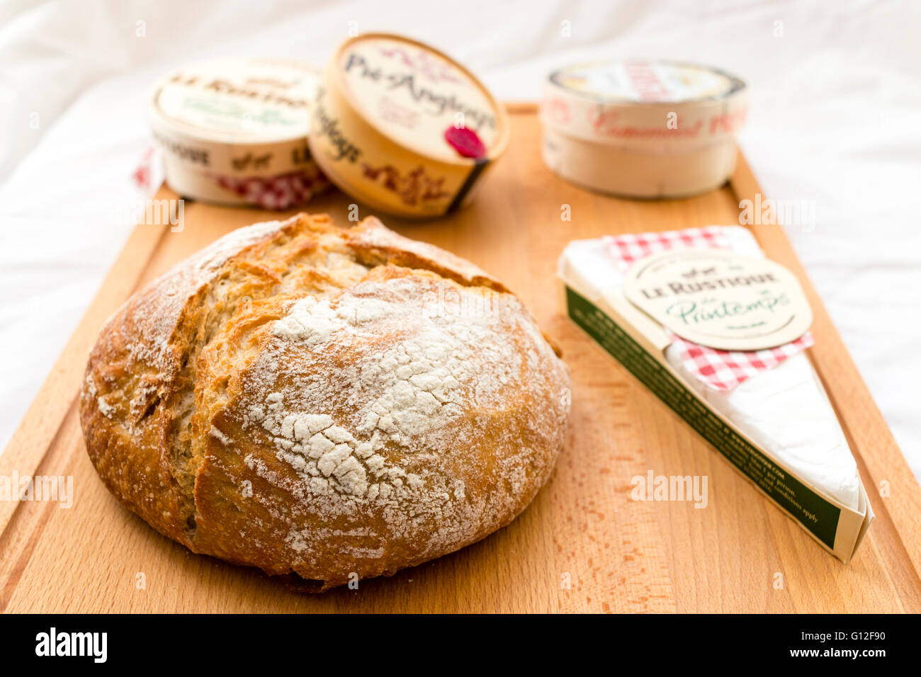 Französischer Käse, Boxed und verpackt, auf Holz Schneidebrett mit runden Sauerteigbrot Laib. Camembert, Brie, verschiedene Marken. Hintergrund mit einem weißen Tuch. Stockfoto