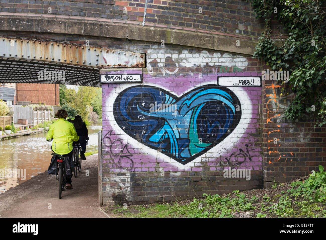 Frauen-Radfahrer, Radfahren auf dem Grand Union Canal Treidelpfad vorbei ein Herz geformt Graffiti-Kunst auf der Brücke Wand, England, UK. Stockfoto