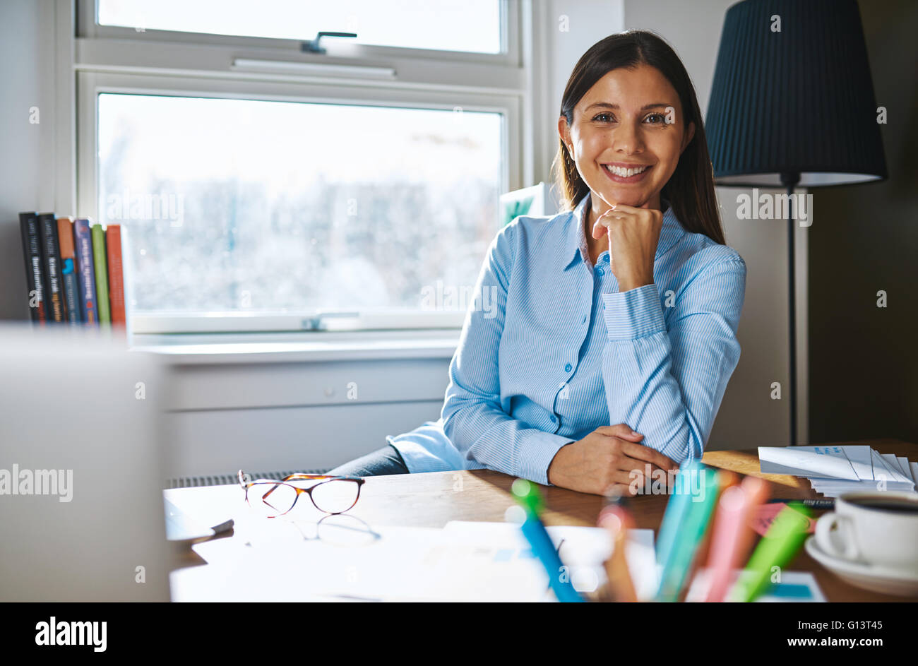 Fröhliche Frau mit der Hand am Kinn trägt blaue Shirt im Sitzen am Schreibtisch bedeckt mit Papieren, Kaffee und Brillen und Fenster i Stockfoto