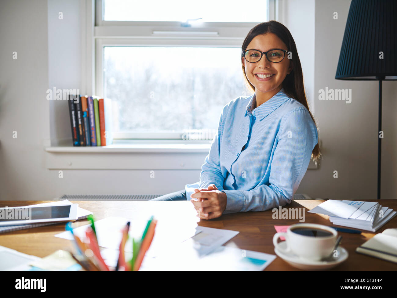 Frau trägt Brille und blaues Hemd am Schreibtisch mit gefalteten Händen neben volle Tasse Kaffee und arbeiten im home Office selbstständig Stockfoto