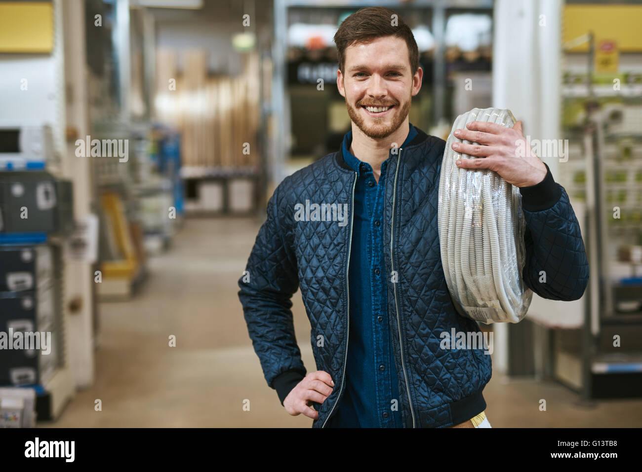 Lächelnde junge Handwerker mit einem Kabel über die Schulter grinsend in die Kamera, als er einen Kauf in einem Baumarkt macht Stockfoto