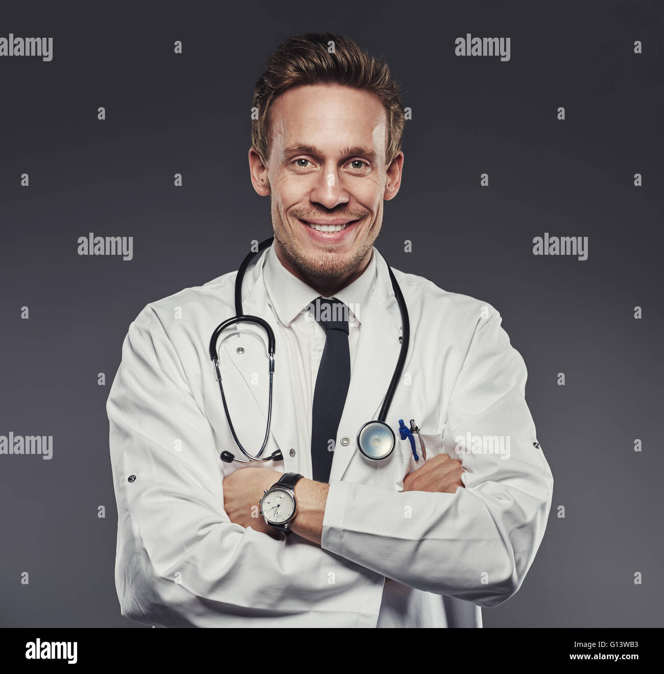 Junge glücklich und freundliche Arzt posiert mit verschränkten Armen während des Tragens medizinische Weißkittel, Armbanduhr und ein Stethoskop, Ill Stockfoto