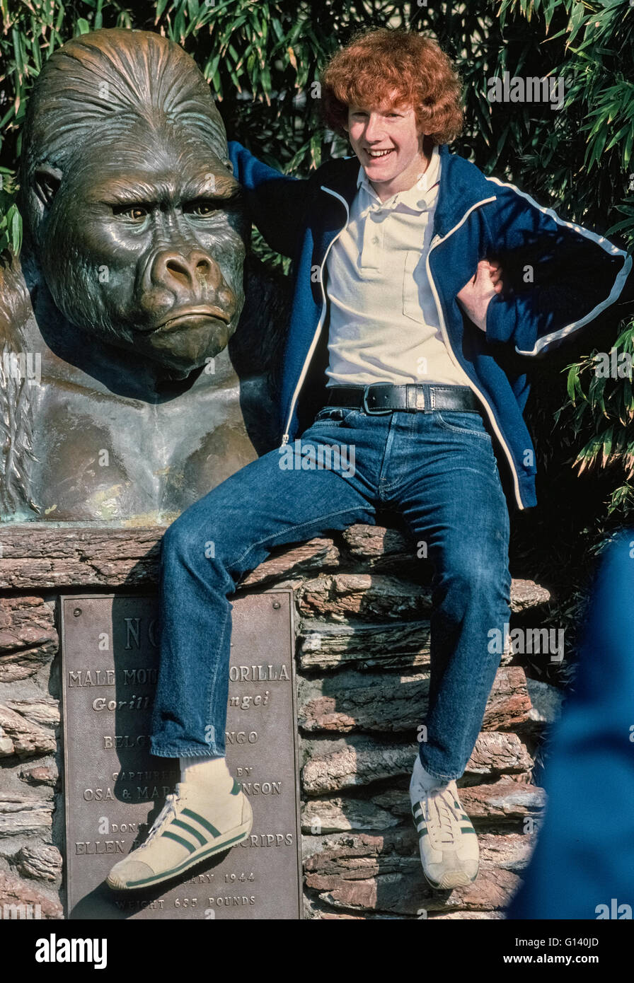 Ein Denkmal mit dem Stirnrunzelnd Gesicht eines männlichen Gorillas aus Afrika erscheint verärgert, die ein Teenager mit einer Hand unter seine Achsel versucht, die Menschenaffen zu imitieren, bei einem Besuch der weltberühmten San Diego Zoo in San Diego, Kalifornien, USA. Die Bronzeskulptur ist eine Hommage an Ngagi, einem östlichen Flachlandgorilla gefangen im damaligen Belgisch-Kongo und in den Zoo in 1931. Die 635-Pfund-Ape starb dort im Jahre 1944. Diese Büste geformten zwei Jahre vor Ngagis Tod war und ist nach wie vor ein beliebter Ort für Bilder in den 100 Hektar großen Zoo zu posieren, die Heimat von mehr als 3.500 seltenen und vom Aussterben bedrohte Tiere. Stockfoto