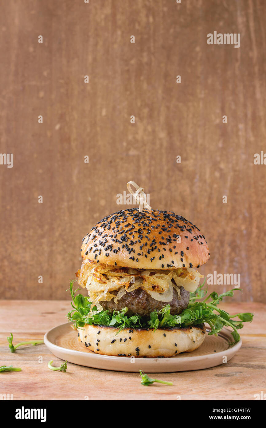 Hausgemachte Burger mit Rindfleisch, gebratenen Zwiebeln und Erbsen-Sprossen, serviert auf Keramikplatte über hölzerne strukturierten Hintergrund Stockfoto