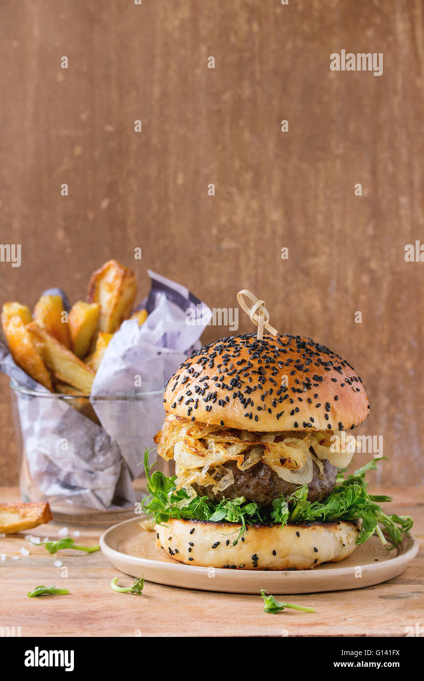 Hausgemachte Burger mit Rindfleisch, gebratenen Zwiebeln und Erbsen-Sprossen, serviert auf Keramikplatte mit Bratkartoffeln über hölzerne strukturierte backgr Stockfoto