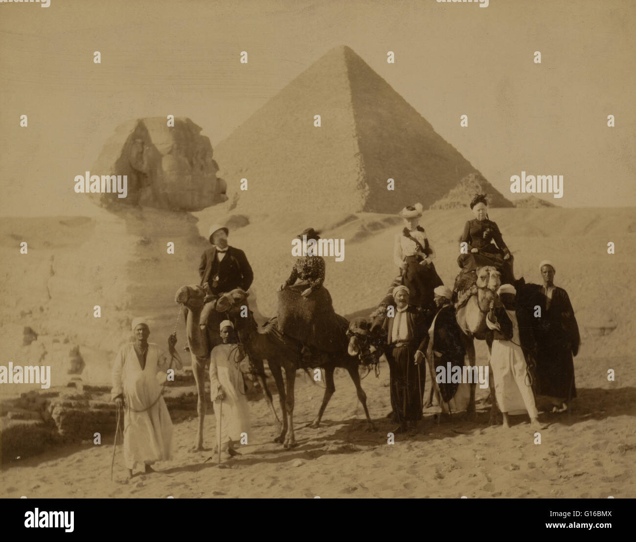 Unter dem Titel: "drei Frauen und ein Mann in der westlichen Kleidung, sitzen auf Kamelen, stehen mehrere einheimische Männer vor die Kamele, die Zügel, eine Pyramide und den Sphinx im Hintergrund". Die Nekropole von Gizeh (Pyramiden von Gizeh) ist eine archäologische Stätte auf dem Gizeh-Plateau Stockfoto