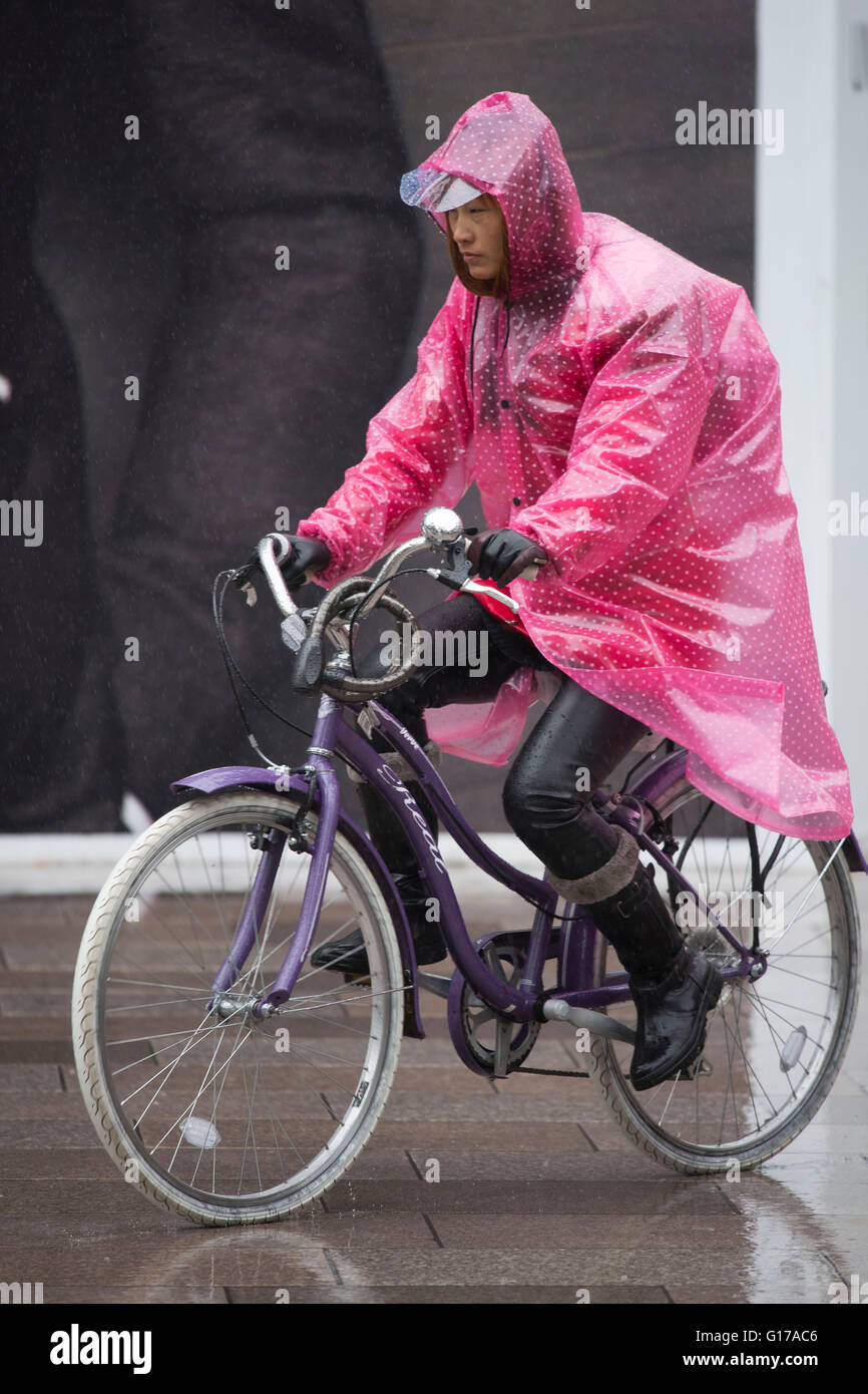 Eine asiatische Frau trägt einen rosa Poncho während der Fahrt ein Fahrrad bei nassem Wetter in Cardiff, Wales. Stockfoto