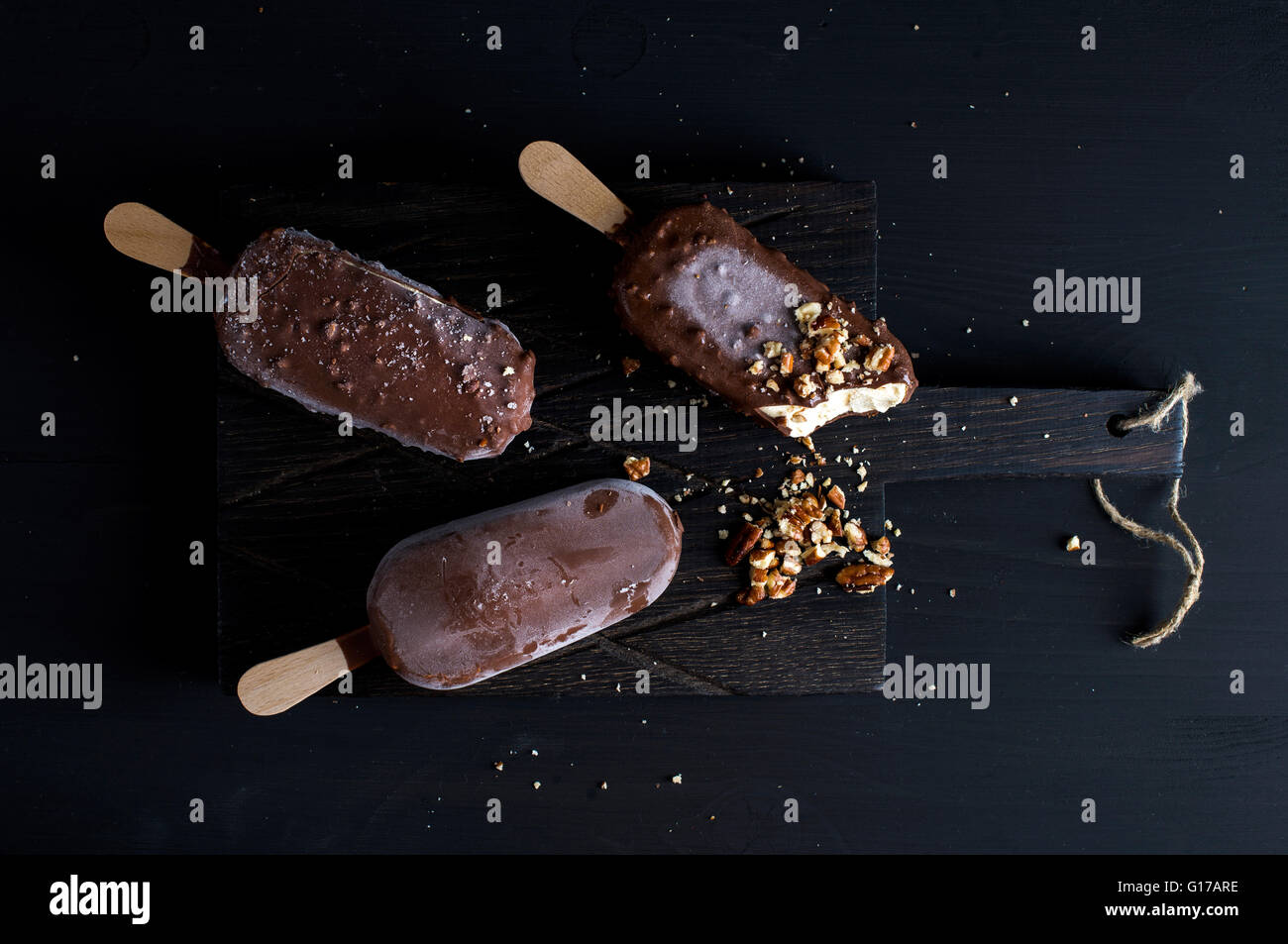 Schokolade getauchte Eis am Stiel mit abgebrochenen Muttern auf dunklem Holz an Bord auf schwarzem Hintergrund, Ansicht von oben, horizontal Stockfoto